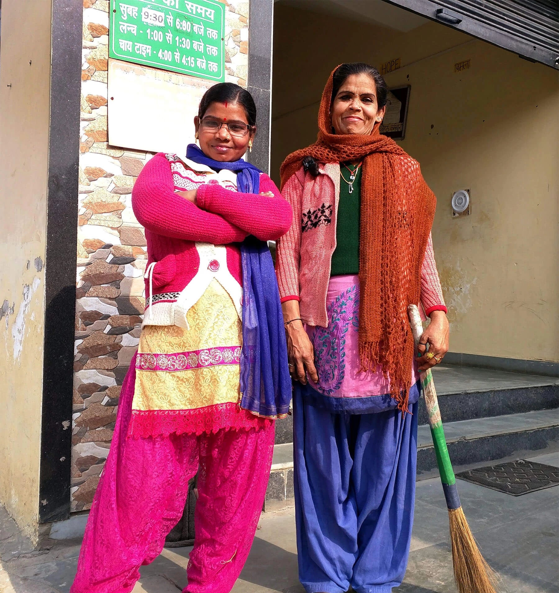 2 Indian women smiling in brightly colored and layered clothing. One is holding a broom.